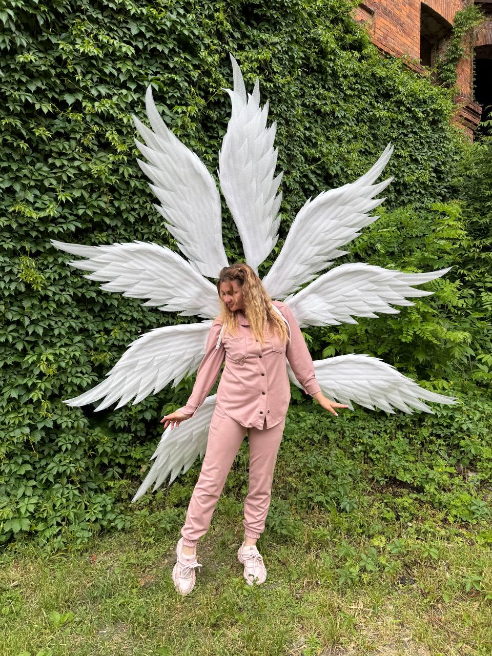 May include: A person wearing a pink outfit and white sneakers stands in front of a wall of green foliage, with large white angel wings attached to their back. The wings are made of multiple layers of feathers.