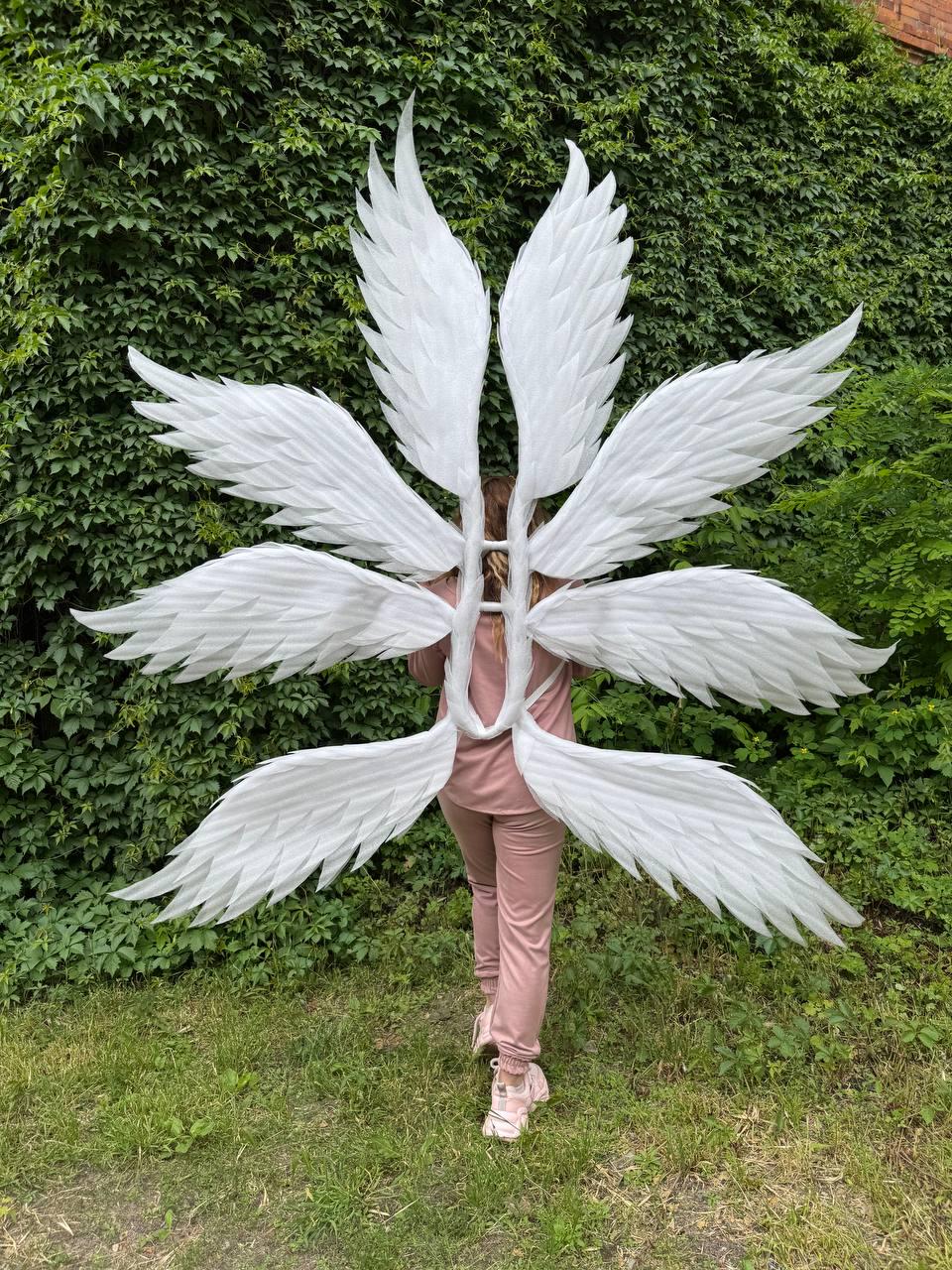 May include: Large, white angel wings with multiple layers of feathers. The wings are attached to a frame worn on the back. The background features green foliage and grass.