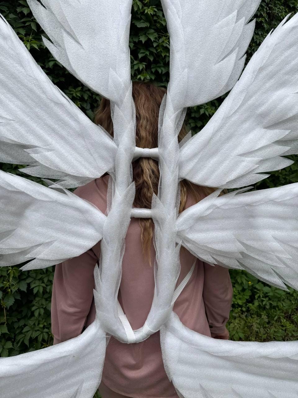 May include: White angel wings with multiple layers of feathers are displayed against a backdrop of green foliage. The wings are attached to a white frame and are worn on the back. The person wearing the wings is wearing a pink sweatshirt.