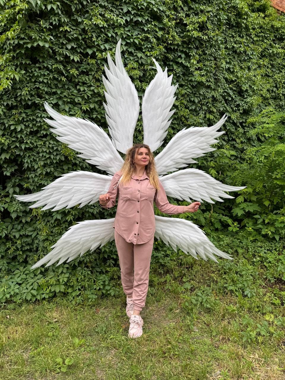 May include: A woman wearing a pink outfit stands in front of a large set of white angel wings. The wings are spread out, with multiple layers of feathers. The background is a wall of green foliage and grass.