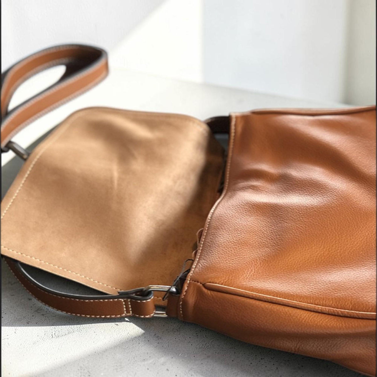 May include: A brown leather shoulder bag with a light brown interior. The bag has a long, adjustable strap and a silver-colored buckle. The leather has a smooth texture and the stitching is white.