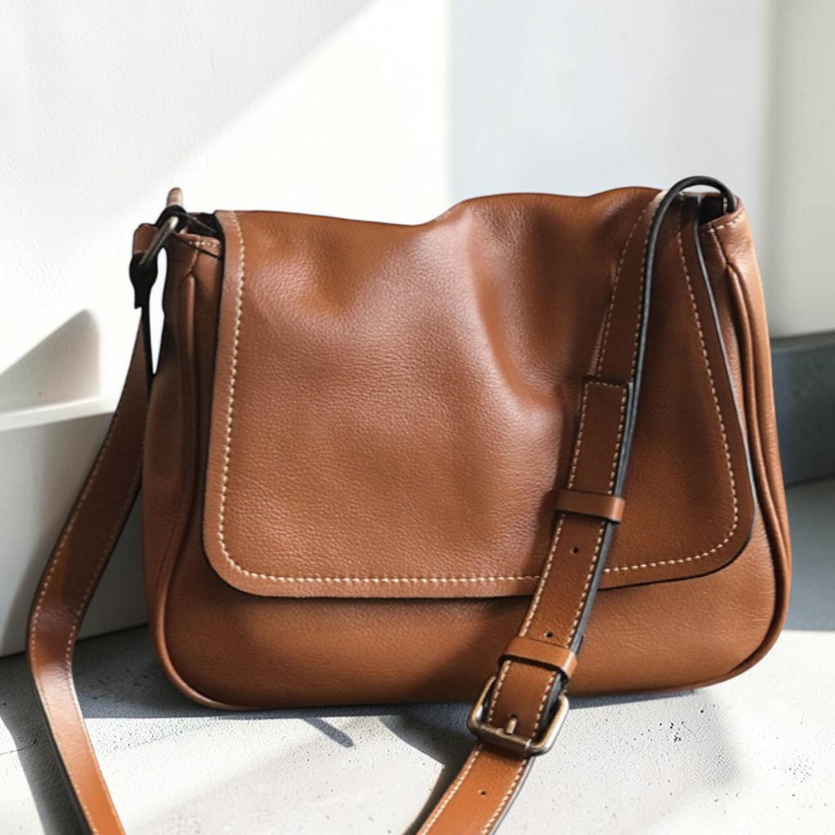 May include: A brown leather crossbody bag with a flap closure and adjustable strap. The bag features white stitching and a metal buckle. The bag is made of a soft, textured leather.