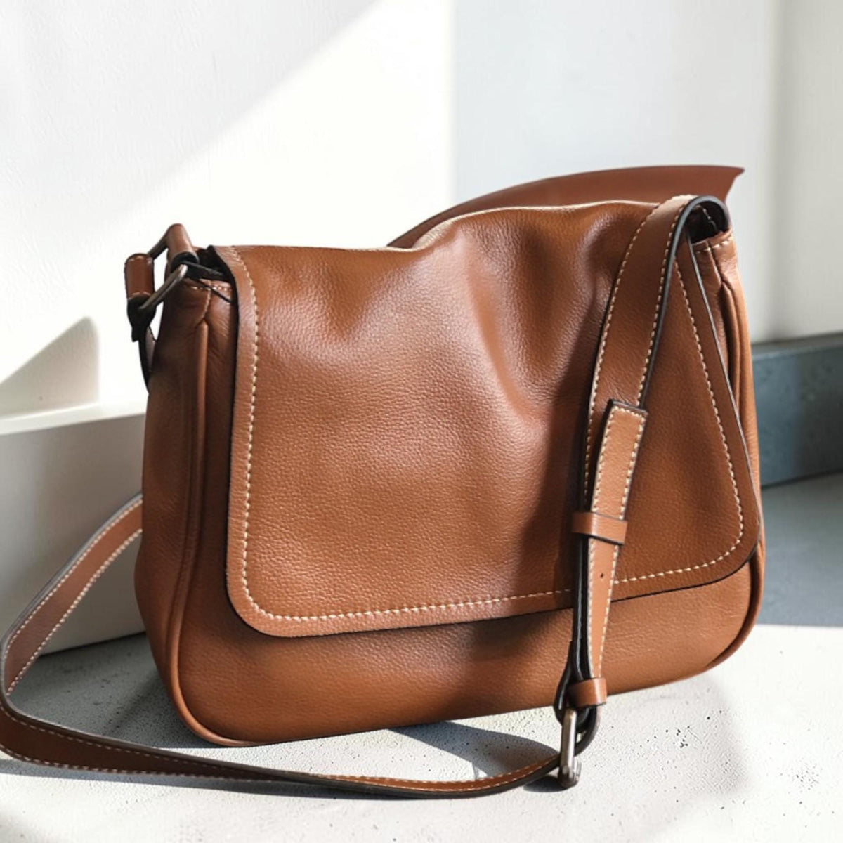 May include: A brown leather messenger bag with a flap closure and adjustable shoulder strap. The bag features white stitching along the edges and a textured surface. The bag is designed for everyday use.