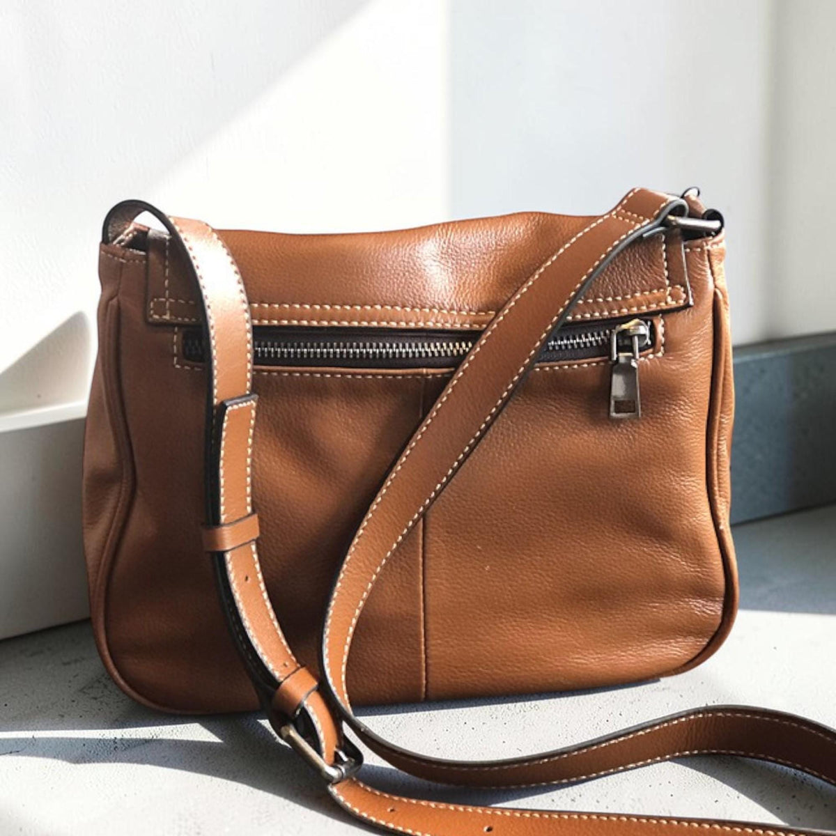 May include: A brown leather crossbody bag with a long adjustable strap. The bag has a zipper closure and a smaller zipper pocket on the front. White stitching accents the edges of the bag and strap. The bag is approximately 10 inches wide and 8 inches tall.