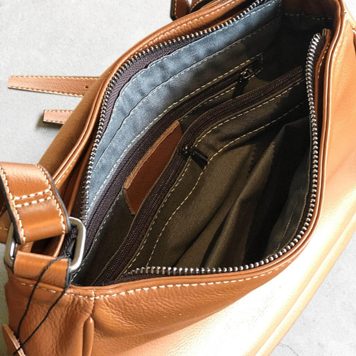 May include: Open view of a brown leather shoulder bag. The bag features multiple zippered compartments with gray and brown fabric lining. Visible are the bag's straps, stitching, and metal hardware.
