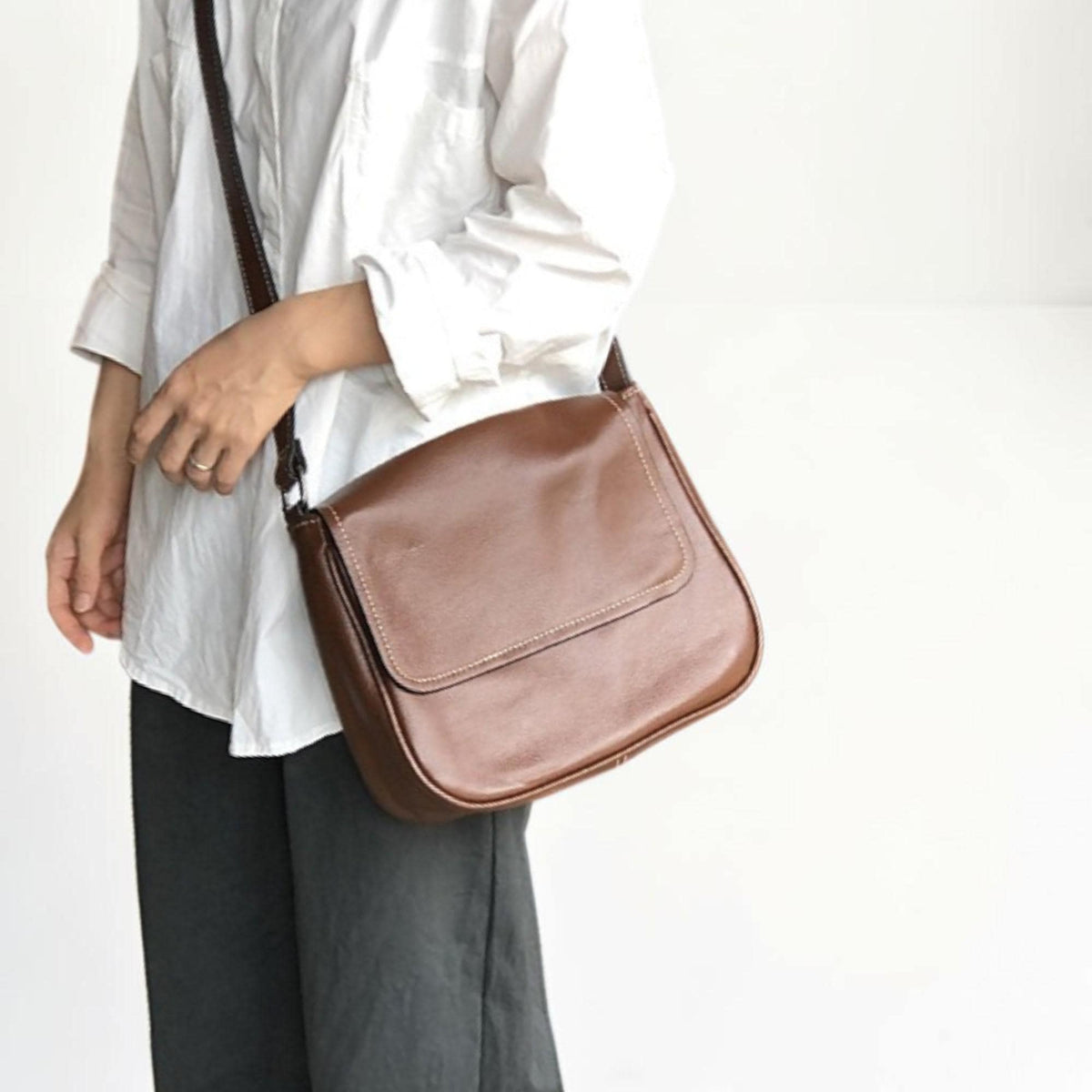 May include: A brown leather messenger bag with a flap closure and a long adjustable strap. The bag has white stitching around the edges and is worn over the shoulder. The bag is made of a smooth, shiny leather.