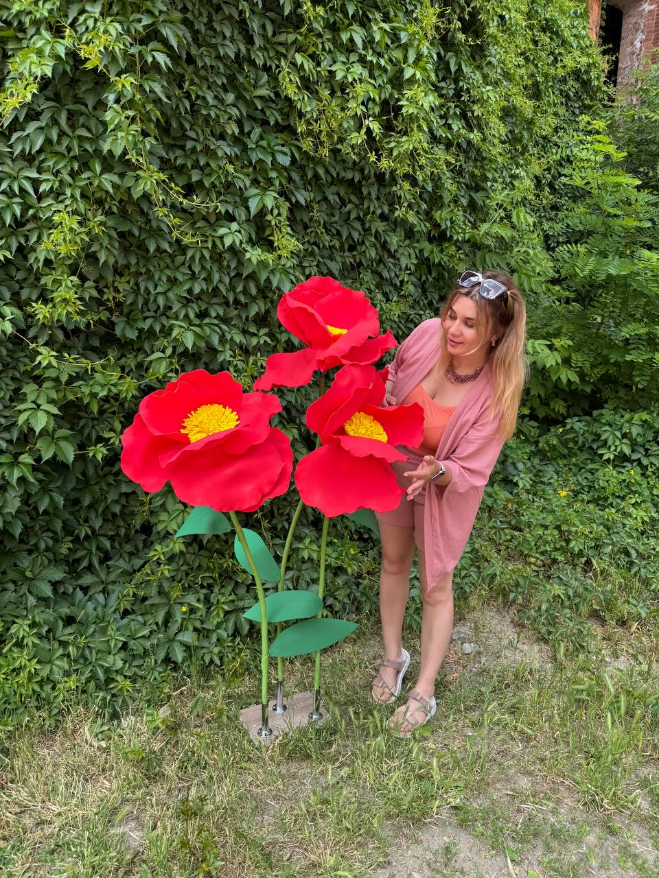 May include: Three oversized artificial red flowers with bright yellow centers, mounted on thin green stems with green leaves, standing on a rectangular base. The flowers feature large, rounded petals and are positioned upright in a vertical arrangement.