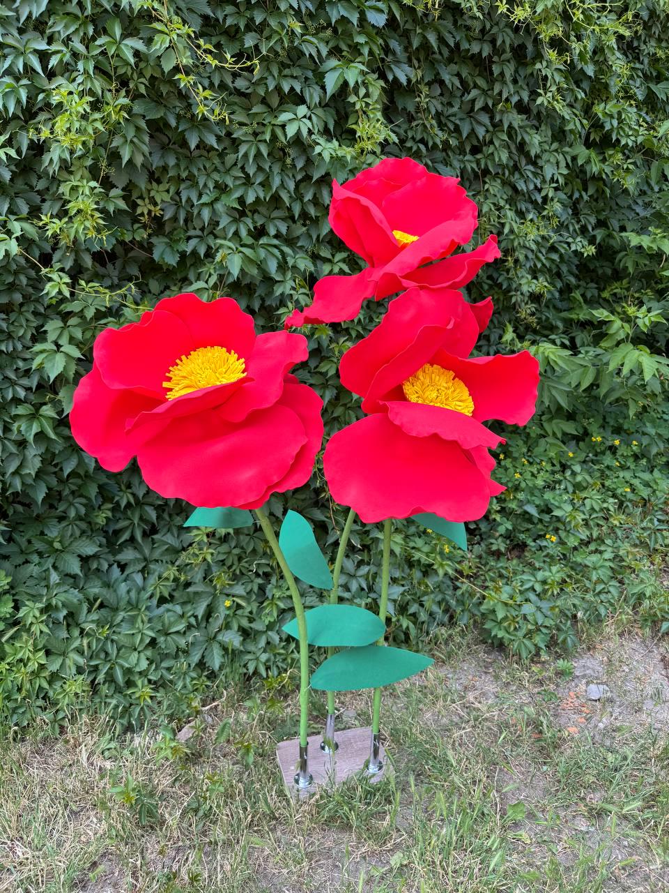 May include: Three large, artificial red flowers with bright yellow centers, each featuring wide, rounded petals. The flowers are attached to thin green stems with green leaves, all supported by a single rectangular base with metal connectors.