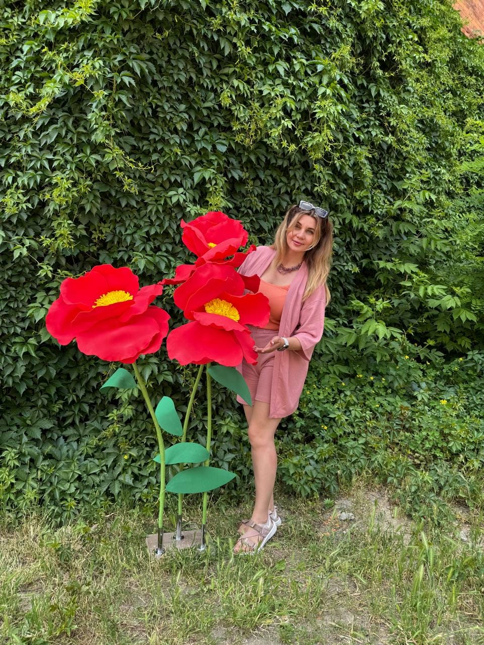 May include: A large decorative floor sculpture featuring three oversized red flowers with bright yellow centers, supported by green stems and leaves on a rectangular base, standing next to a person wearing a pink blazer and shorts set.