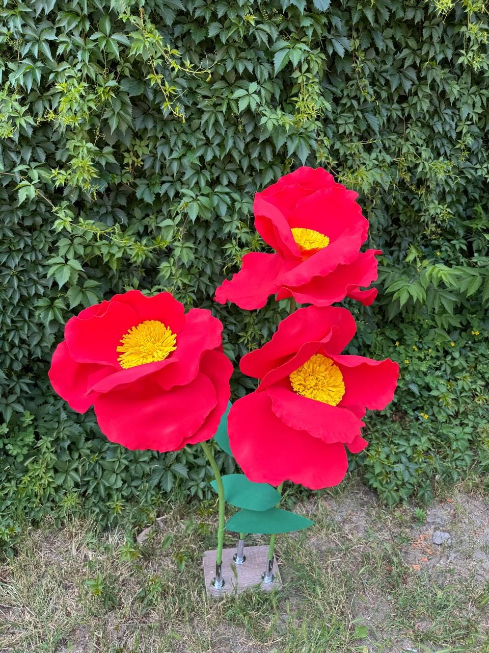 May include: Three large, artificial red flowers with bright yellow centers, each featuring wide, layered petals. The flowers are attached to green stems with green leaves, all mounted on a small, square, light-colored base that supports the stems.