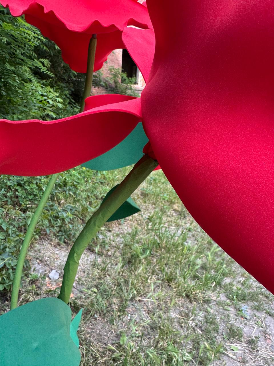 May include: Large artificial red flowers with smooth, rounded petals and green stems. The flowers feature a matte texture and are supported by thin, green stalks with flat, rounded green leaves attached at the base of the stems.