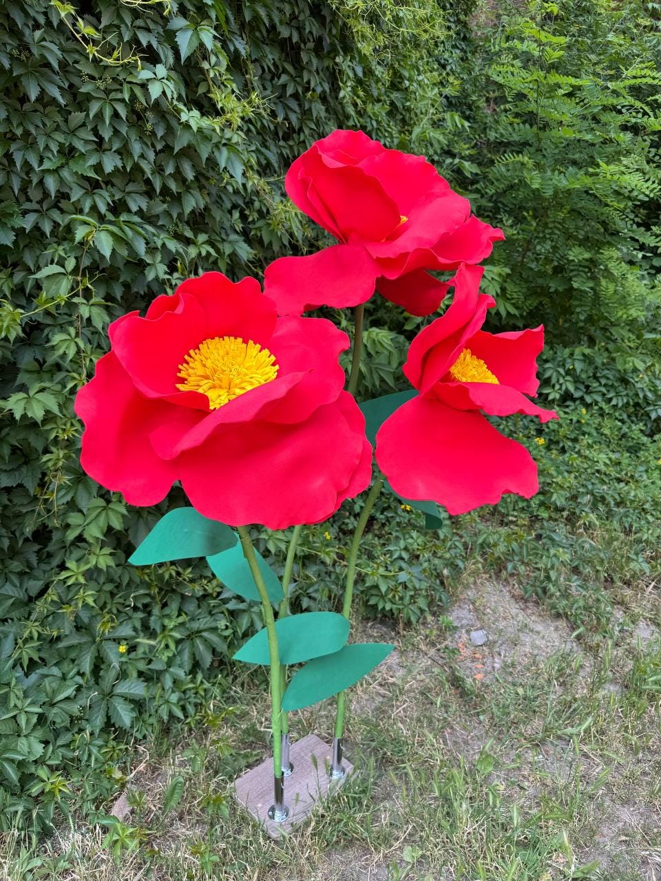 May include: Three large, oversized artificial flowers with vibrant red petals and bright yellow centers. Each flower is attached to a thin green stem with green leaves, and all three stems are mounted into a rectangular base with metal supports.
