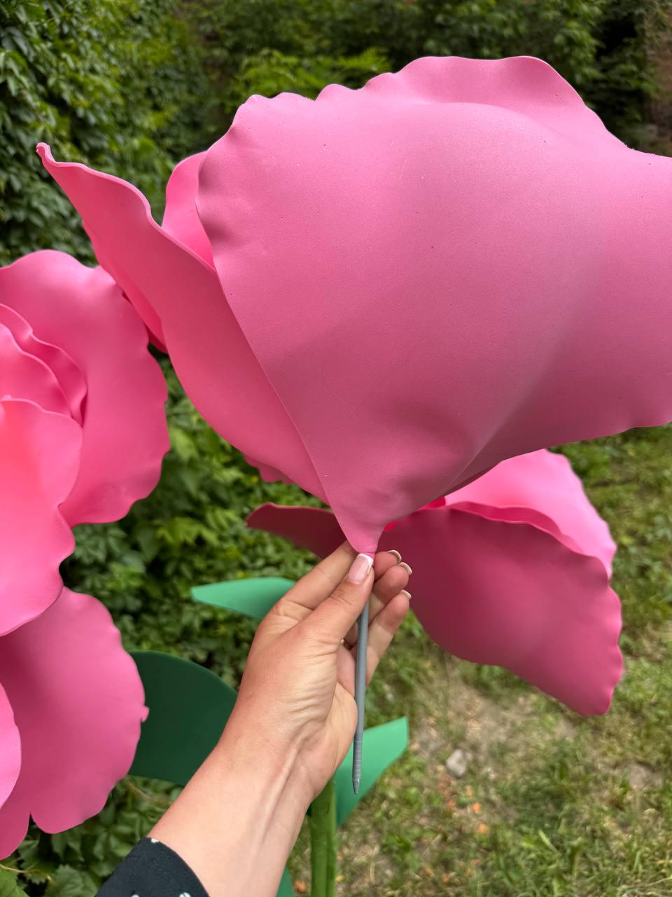 May include: A large, oversized artificial pink flower with smooth, rounded petals attached to a thin, straight gray stem. The flower features a vibrant, solid pink color throughout its petals, which are shaped to mimic a blooming rose or peony.