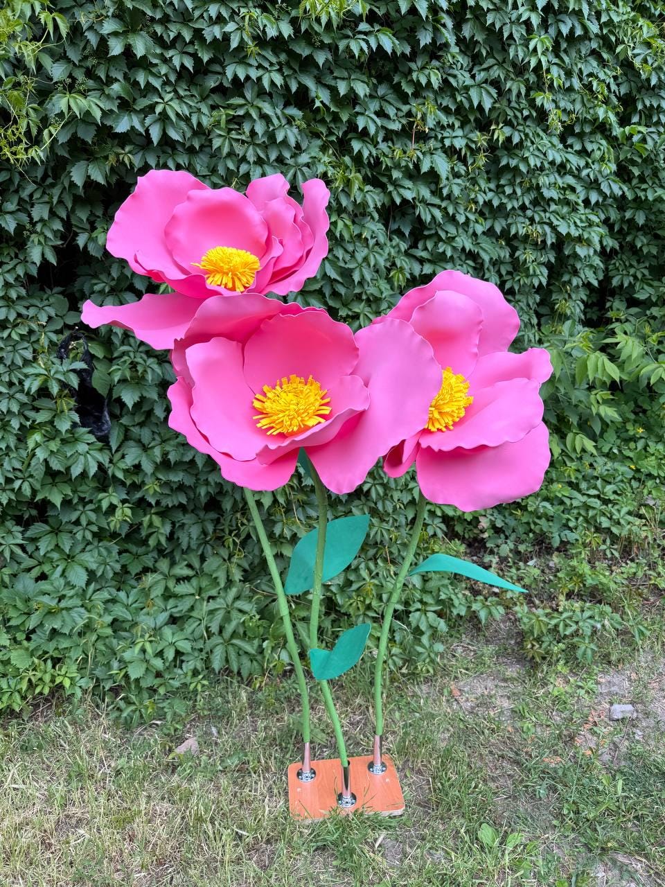 May include: Three large, artificial pink flowers with bright yellow centers, each featuring layered, rounded petals. The flowers are attached to thin green stems with green leaves, all mounted on a rectangular wooden base with metal connectors.