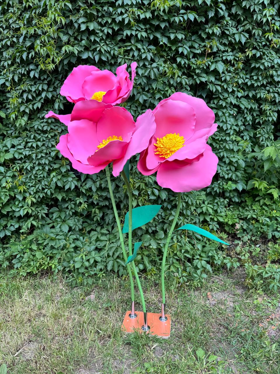 May include: Three large, artificial pink flowers with bright yellow centers, each mounted on a thin green stem. The stems are attached to a rectangular orange base with silver metal connectors, creating a freestanding decorative floral arrangement.