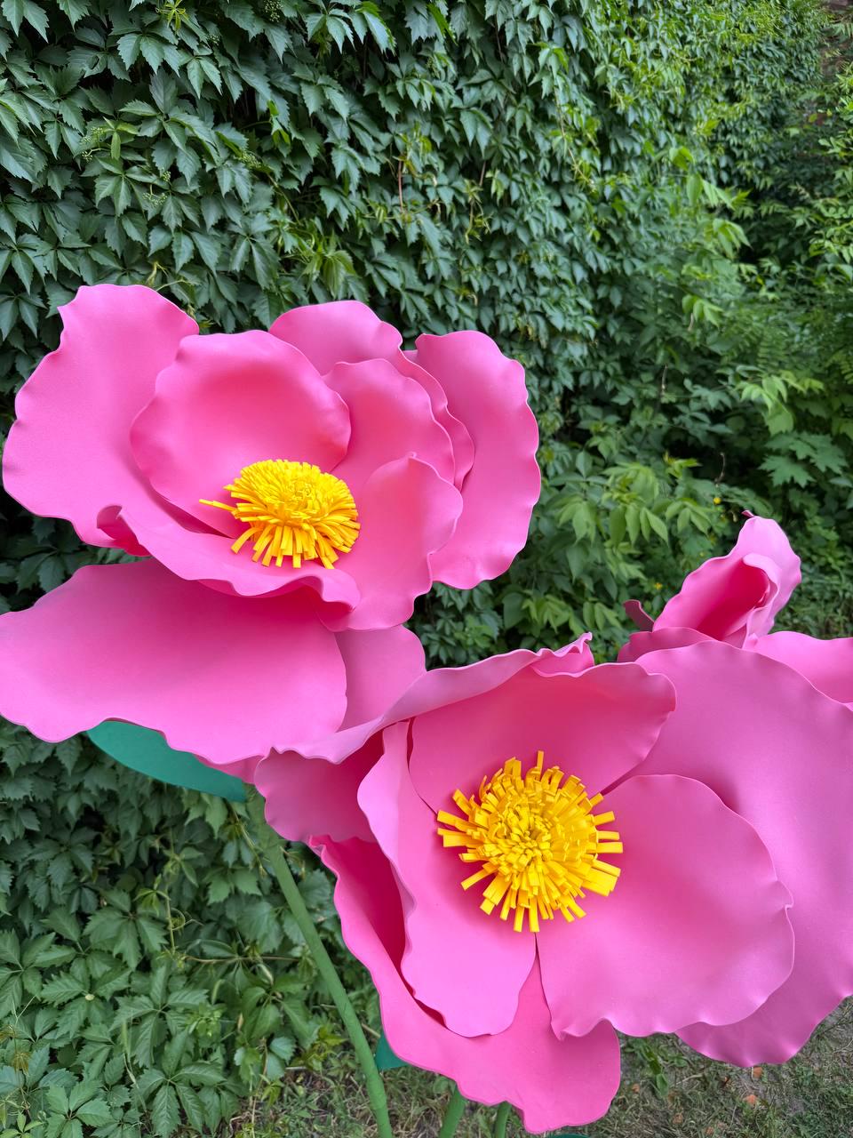 May include: Two large, oversized artificial flowers with vibrant pink, smooth, rounded petals and a bright yellow, fringed, textured center. The flowers are mounted on thin green stems, creating a bold, sculptural display of floral art.