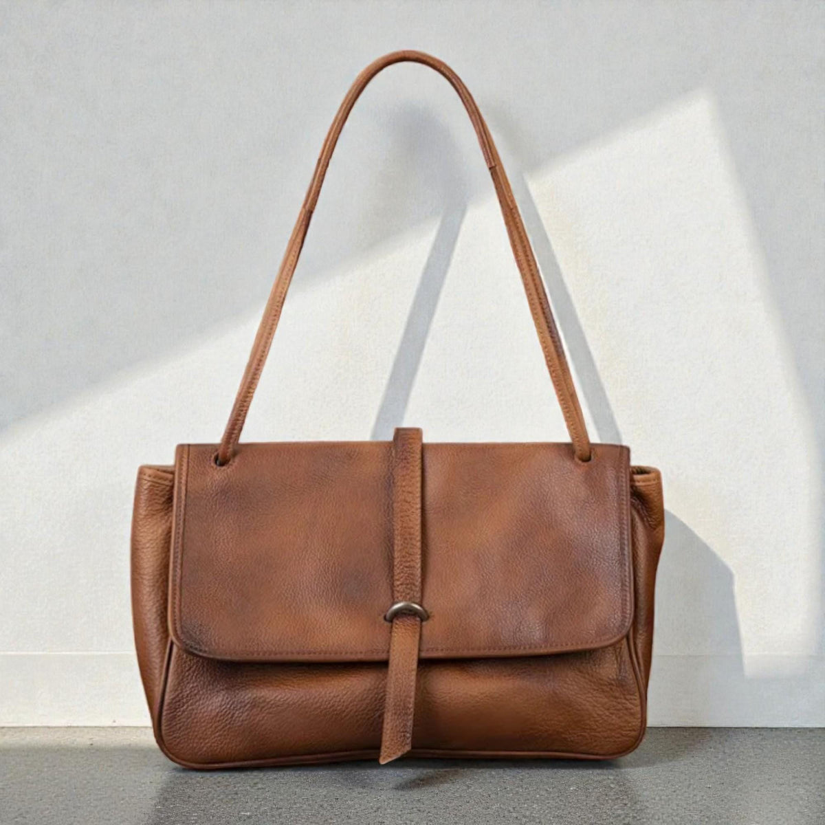 May include: A brown leather shoulder bag with a flap closure and a long strap. The bag has a rectangular shape with a textured surface and a strap that secures the flap. The bag is set against a white wall.