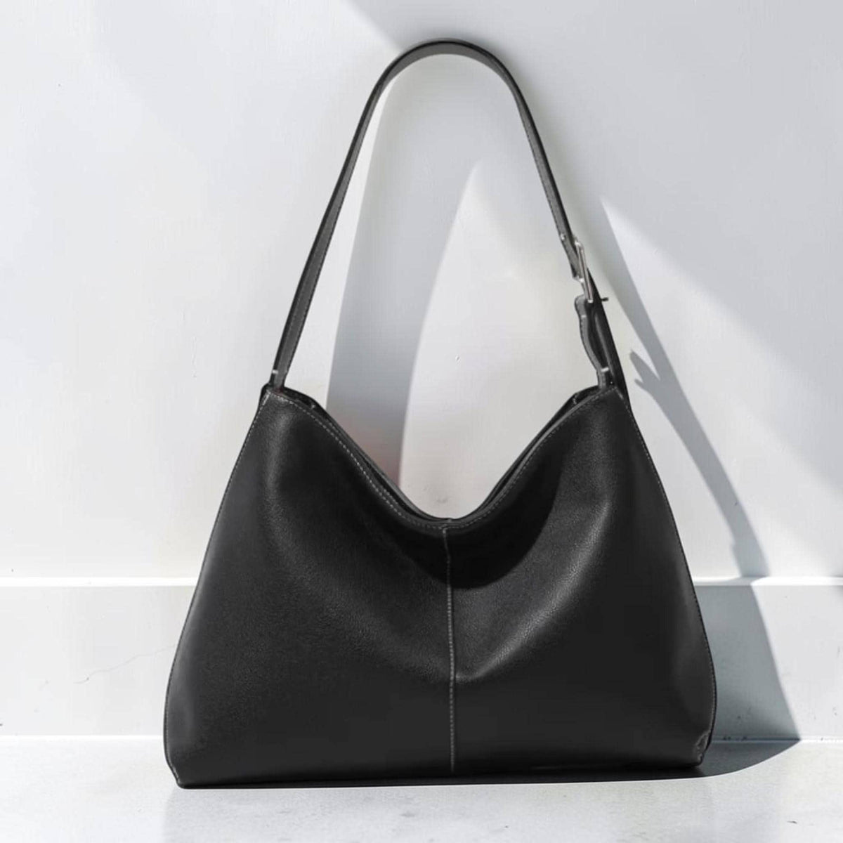 May include: A black hobo-style shoulder bag with a smooth matte finish, featuring a single adjustable strap with a silver-toned metal buckle, a central vertical seam detail, and a curved top opening that creates a soft, slouchy silhouette.
