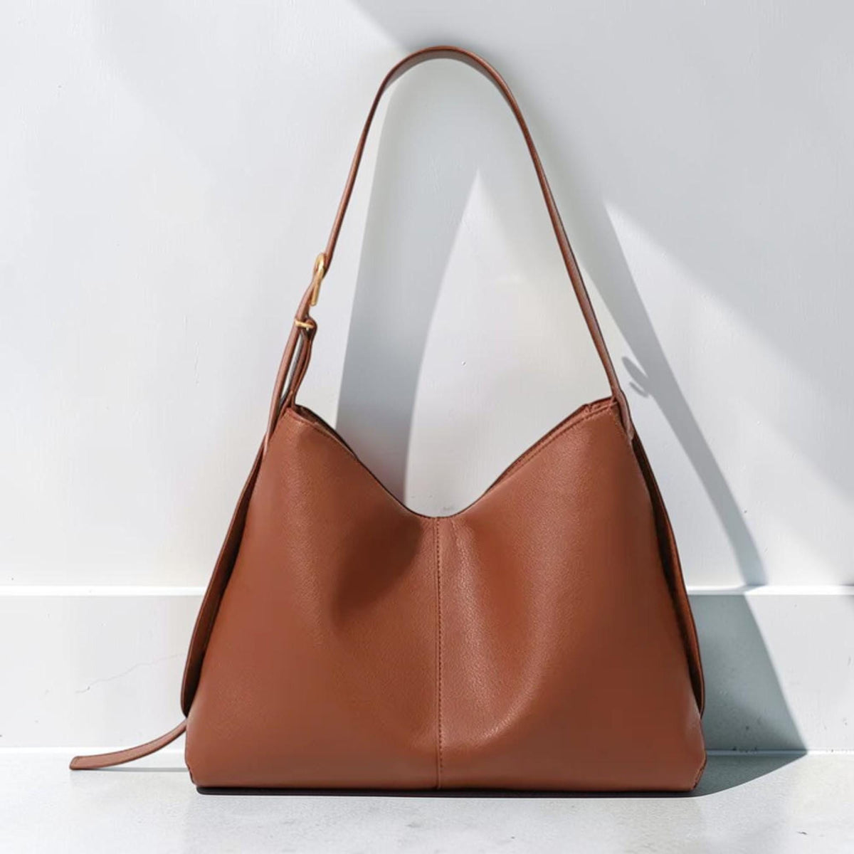 May include: A solid brown hobo-style shoulder bag with a smooth, matte finish. The bag features a single adjustable strap with a gold-toned metal buckle, a central vertical seam, and a wide, curved opening that tapers toward the top handle attachment.