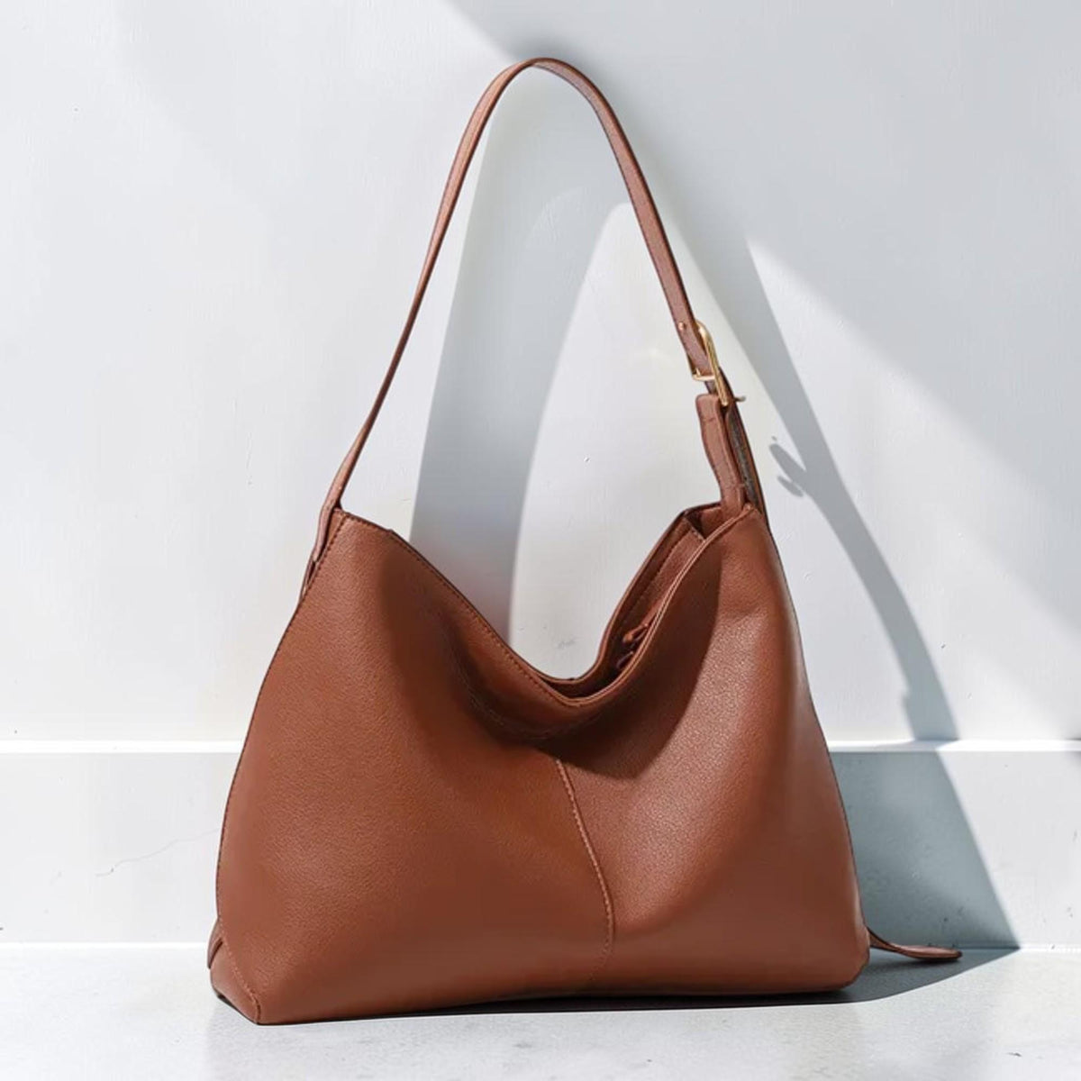 May include: A medium-sized hobo-style shoulder bag in a solid tan-brown color. The bag features a single adjustable strap with a gold-toned metal buckle, a smooth exterior surface, and a central vertical seam detail on the front panel.