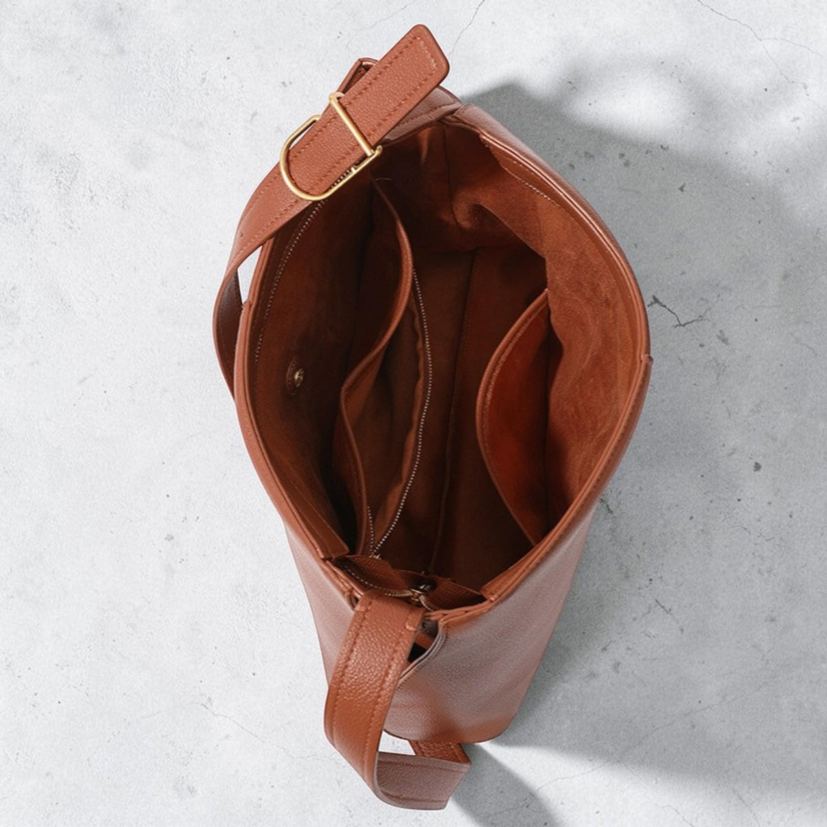May include: A cognac-colored hobo bag with a textured exterior and a smooth interior lining. The bag features an adjustable shoulder strap with a gold-toned metal buckle, an open top, and internal pockets including a zippered compartment.