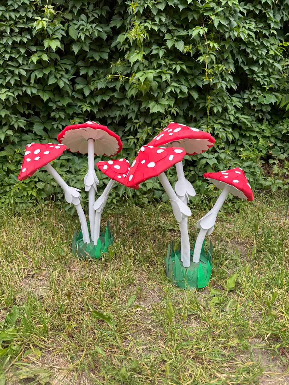 May include: A collection of five artificial mushroom sculptures featuring bright red caps with white polka dots and white stems. Each mushroom is supported by a green base designed to resemble grass, creating a cluster of decorative fungi.