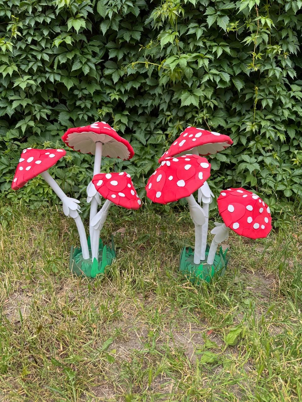 May include: Two clusters of artificial mushrooms featuring bright red caps with white spots. Each mushroom has a slender white stem and a green base. The caps are rounded and vary in size, with some angled outward and others standing upright.