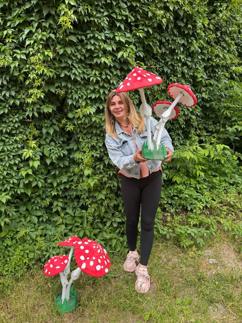 May include: Two sets of decorative artificial mushrooms featuring bright red caps with white spots and white stems. One set is held in the hands, while the other stands on the ground. Each cluster is mounted on a green base resembling grass.