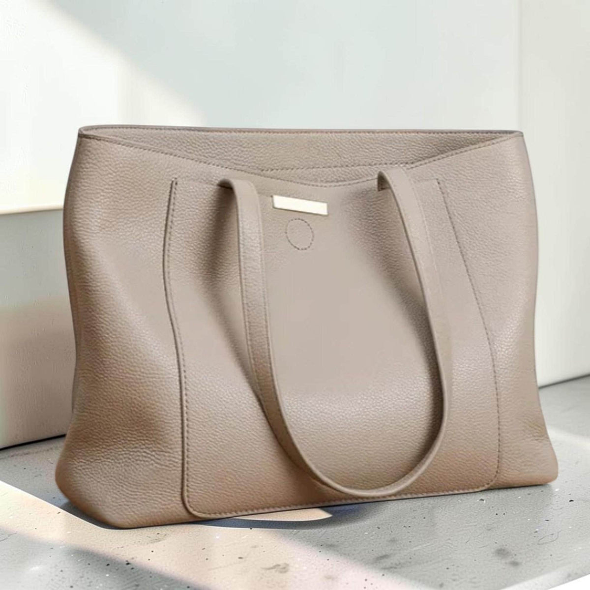 May include: A taupe-colored tote bag with a pebbled leather texture, featuring two shoulder straps, a rectangular front pocket, and a small metallic rectangular accent piece centered near the top opening of the bag.