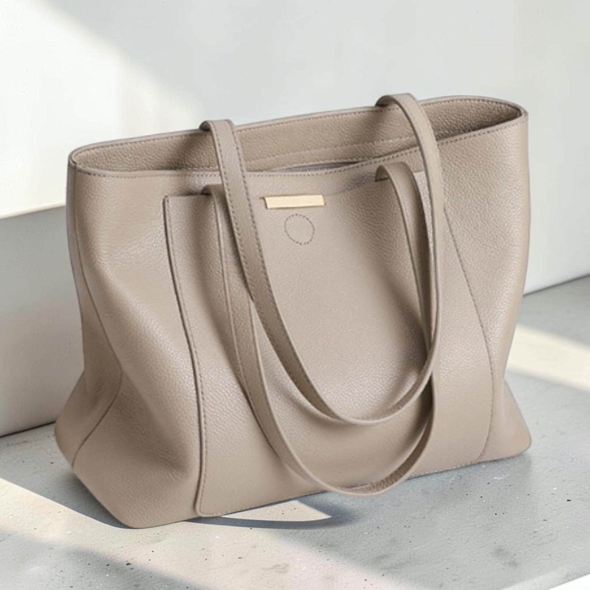 May include: A taupe-colored tote bag with a pebbled texture, featuring two long shoulder straps and a rectangular gold-toned metal accent bar centered on the upper front panel. The bag has a structured base and a clean, open-top design.