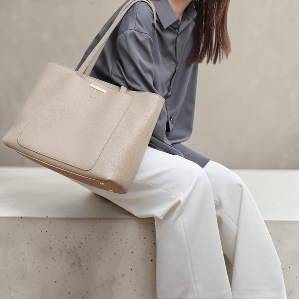 May include: A beige structured tote bag with a smooth, matte finish and a rectangular front pocket. The bag features two thin shoulder straps and a small, horizontal gold-toned metal bar accent located at the top center of the front panel.
