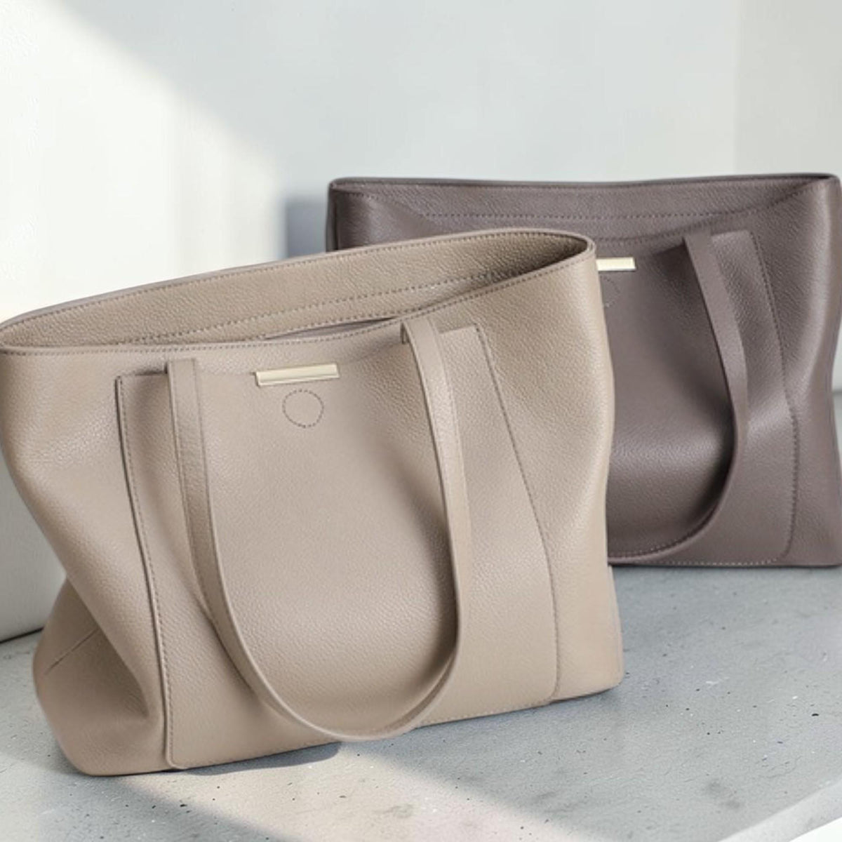May include: Two structured tote bags with a pebbled texture. The front bag is light beige, and the back bag is a darker taupe. Both feature dual shoulder straps, a horizontal metallic bar accent at the top center, and a circular stitched detail.