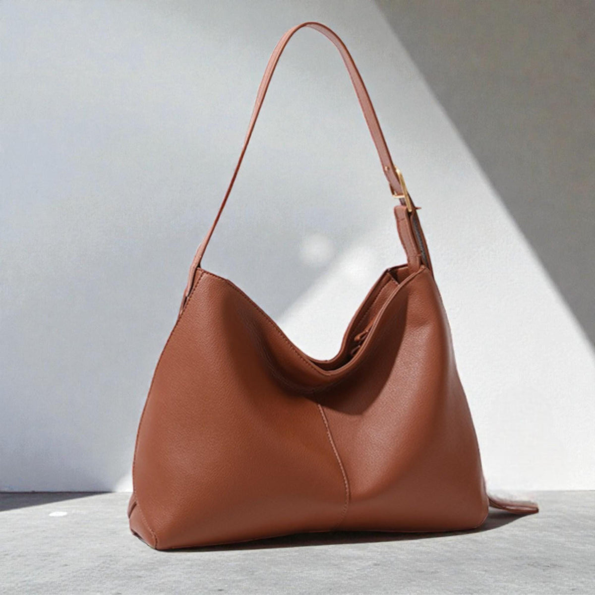 May include: A brown leather shoulder bag with a single strap and a gold-coloured buckle. The bag has a curved shape and a spacious interior. The bag is sitting on a grey surface with a white background.