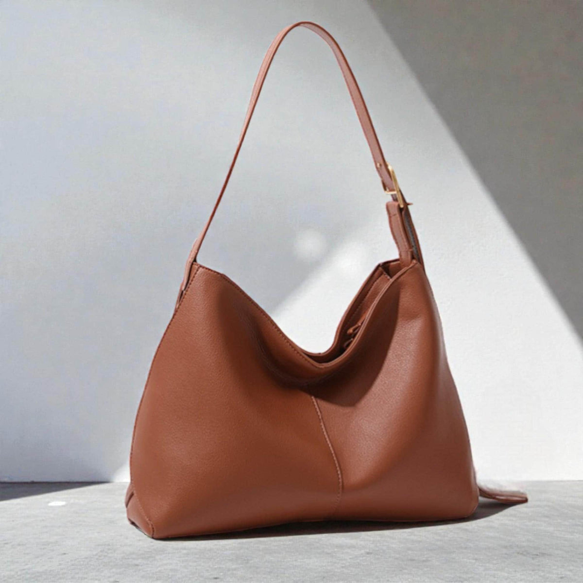 May include: A brown leather shoulder bag with a single strap and a gold-coloured buckle. The bag has a curved shape and a spacious interior. The bag is sitting on a grey surface with a white background.