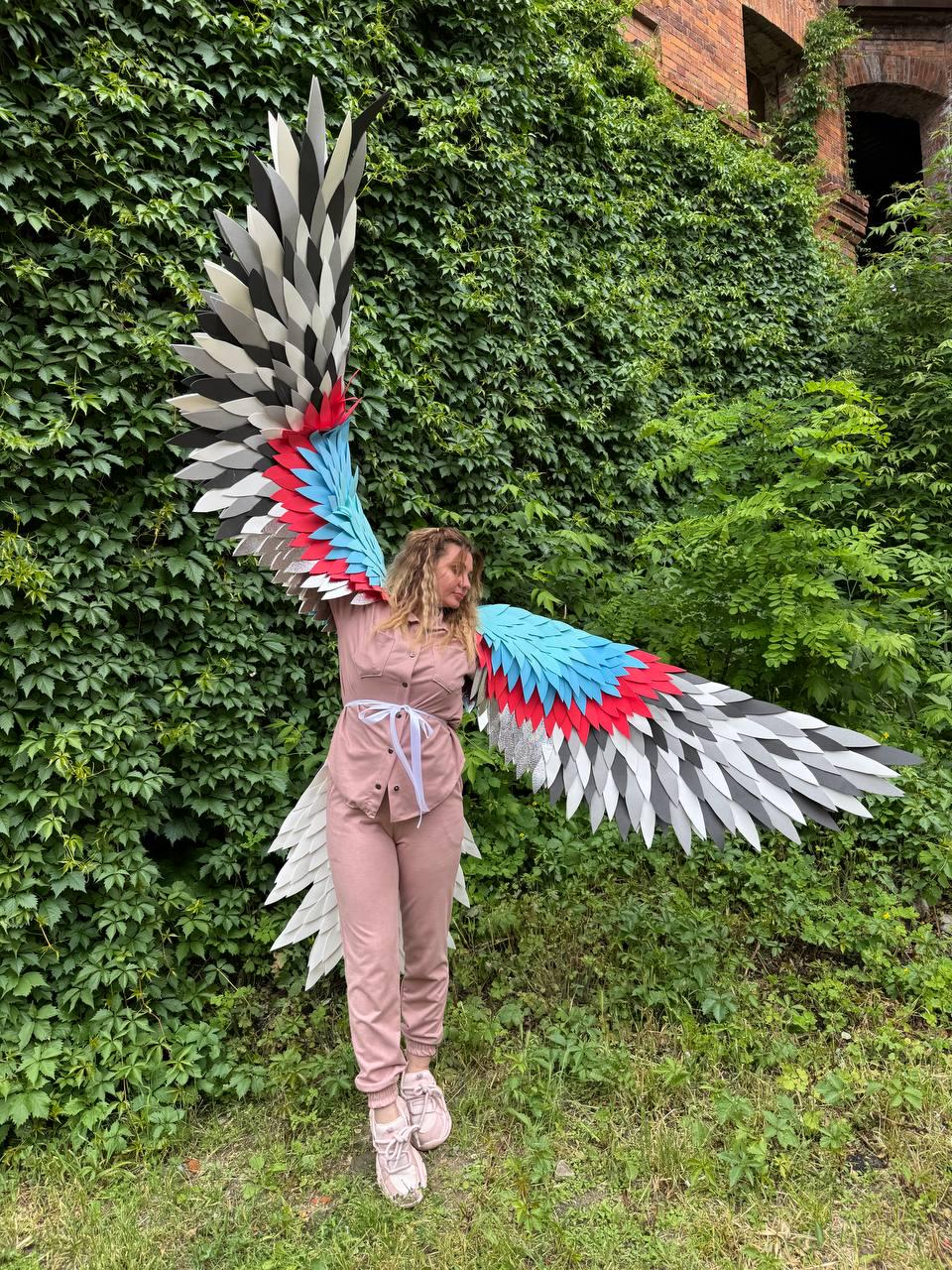 May include: Large decorative wings featuring layered, pointed feathers in a color palette of black, white, gray, bright blue, and red. The wings are attached to a long-sleeved, button-down shirt and matching pants in a solid dusty pink color.