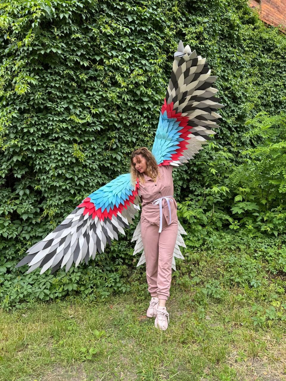 May include: Large decorative wings featuring layered, pointed feathers in a gradient of light blue, bright red, dark gray, and white. The wings are attached to a matching harness worn over a dusty pink button-down shirt and jogger pants set.