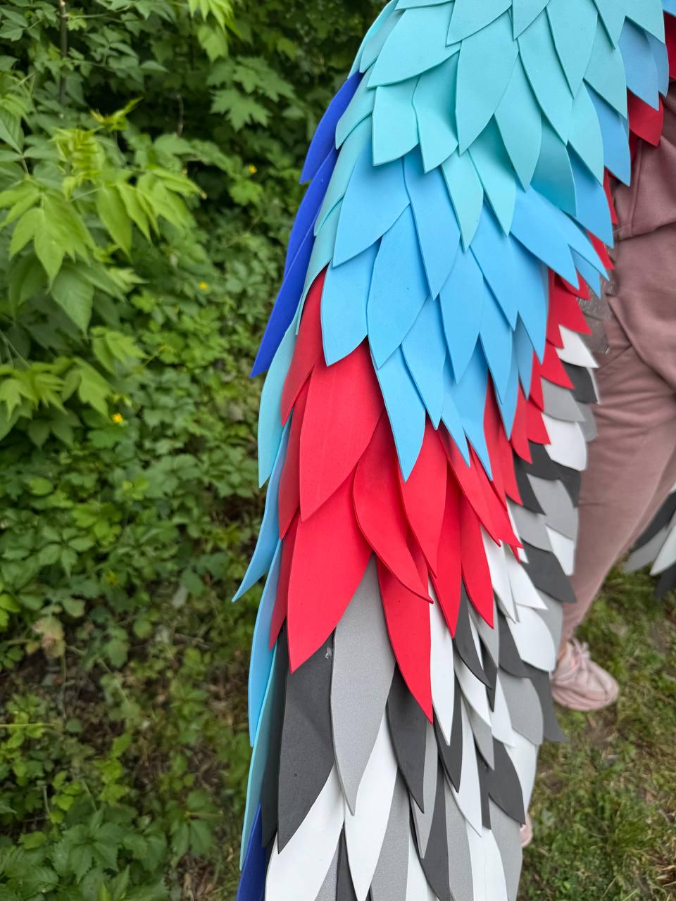 May include: A costume wing accessory featuring layered, leaf-shaped foam feathers in a vibrant color palette of bright blue, light turquoise, deep red, dark gray, and white. The feathers are arranged in overlapping rows to create a textured effect.