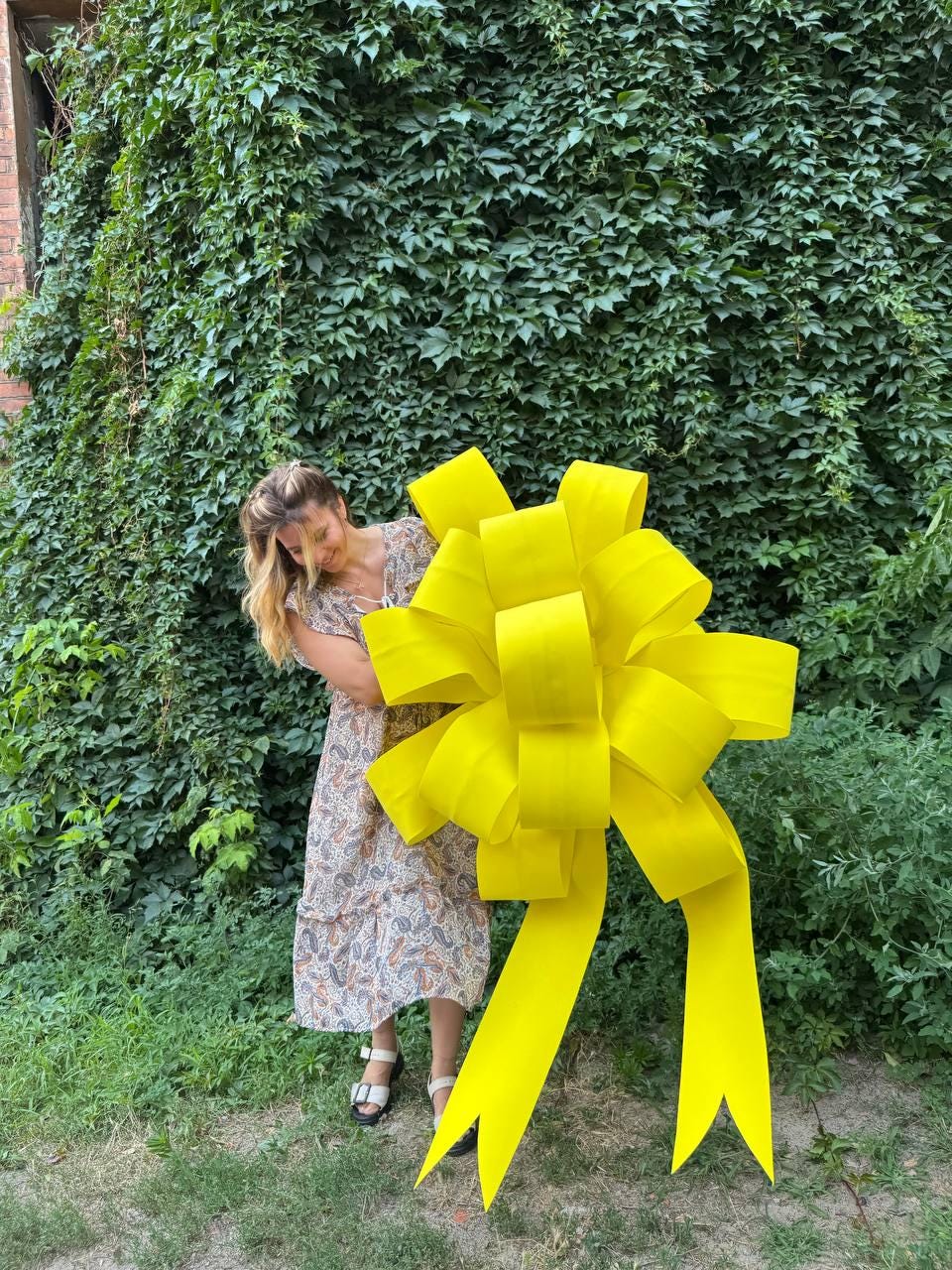 May include: A large, bright yellow decorative bow made of wide ribbon loops, featuring two long, trailing tails with V-shaped notched ends. The bow is constructed from a smooth, solid-colored material and is held in front of a person.