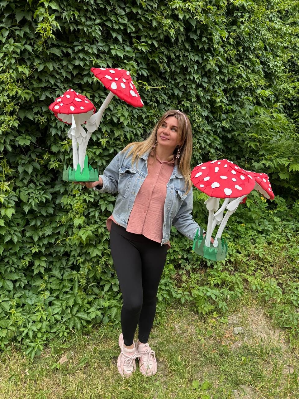 May include: Two large decorative mushroom sculptures featuring bright red caps with white spots, white stems, and green bases. The mushrooms are crafted with a fabric-like texture and are held by a person wearing a denim jacket, pink shirt, and black leggings.