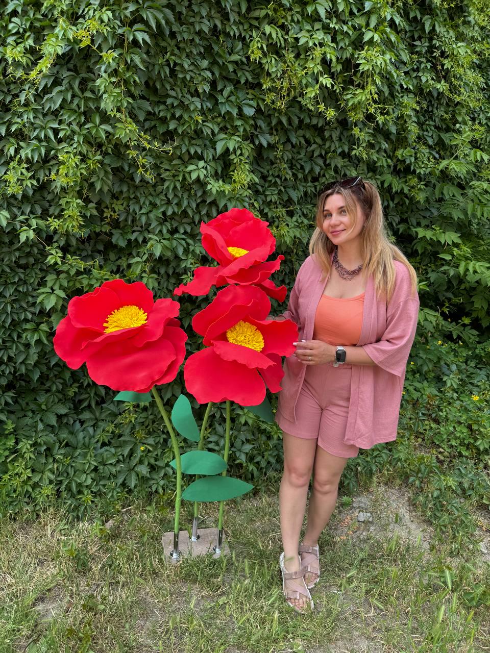 May include: Three oversized artificial red flowers with bright yellow centers, supported by thin green stems and leaves on a small square base, standing next to a person wearing a dusty pink cardigan, matching shorts, and an orange top.