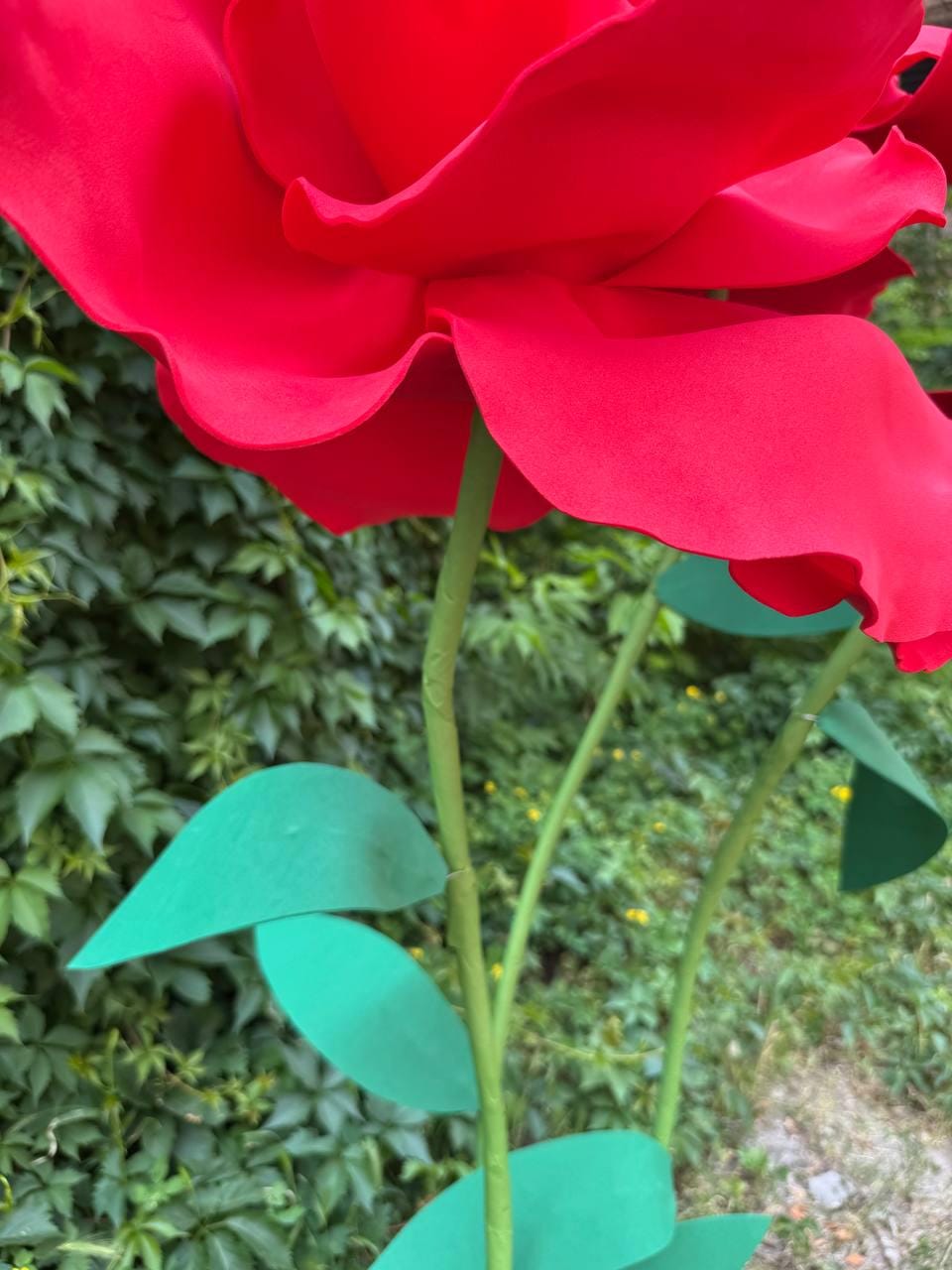 May include: A large, oversized artificial red rose with smooth, curved petals. The flower is attached to a long, slender green stem featuring several flat, pointed green leaves extending from the sides. The structure is rigid and sculptural.
