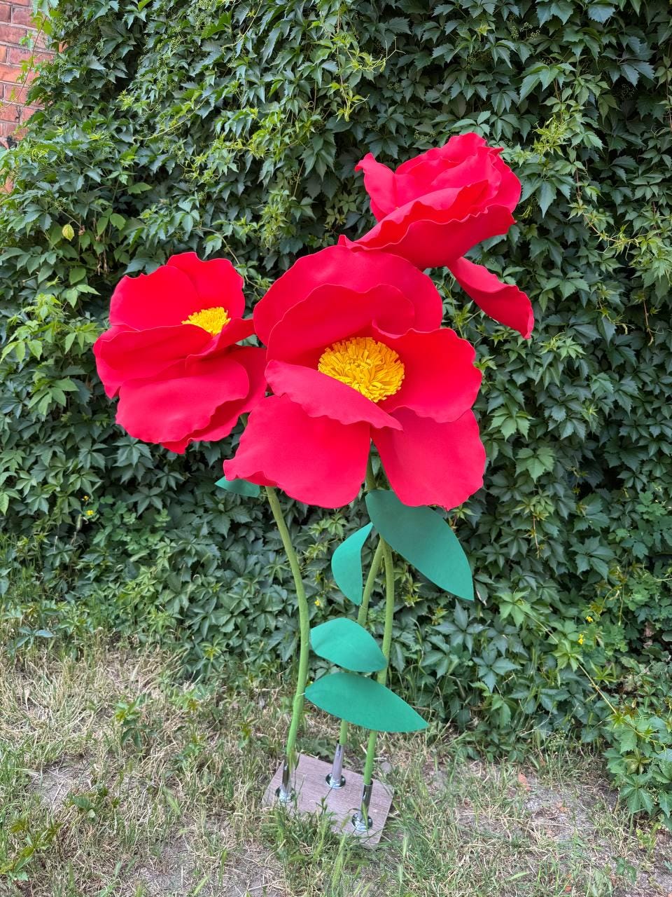 May include: Three large, artificial red flowers with bright yellow centers, each mounted on a thin green stem with green leaves. The flowers are attached to a rectangular base, standing upright with vibrant red petals and textured yellow stamens.