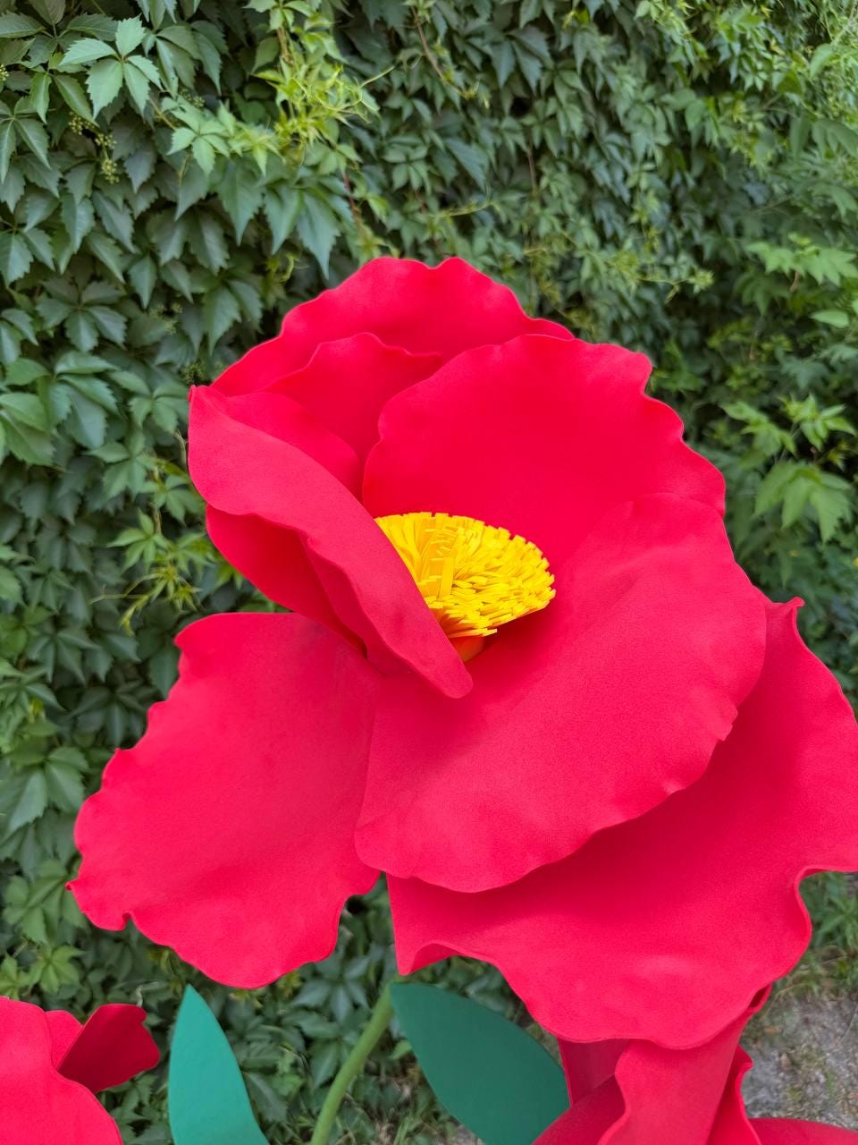 May include: A large, oversized artificial flower with vibrant red, rounded petals and a textured, bright yellow center. The flower features a layered, sculptural design with smooth, matte-finish petals that curve outward, creating a bold, three-dimensional appearance.