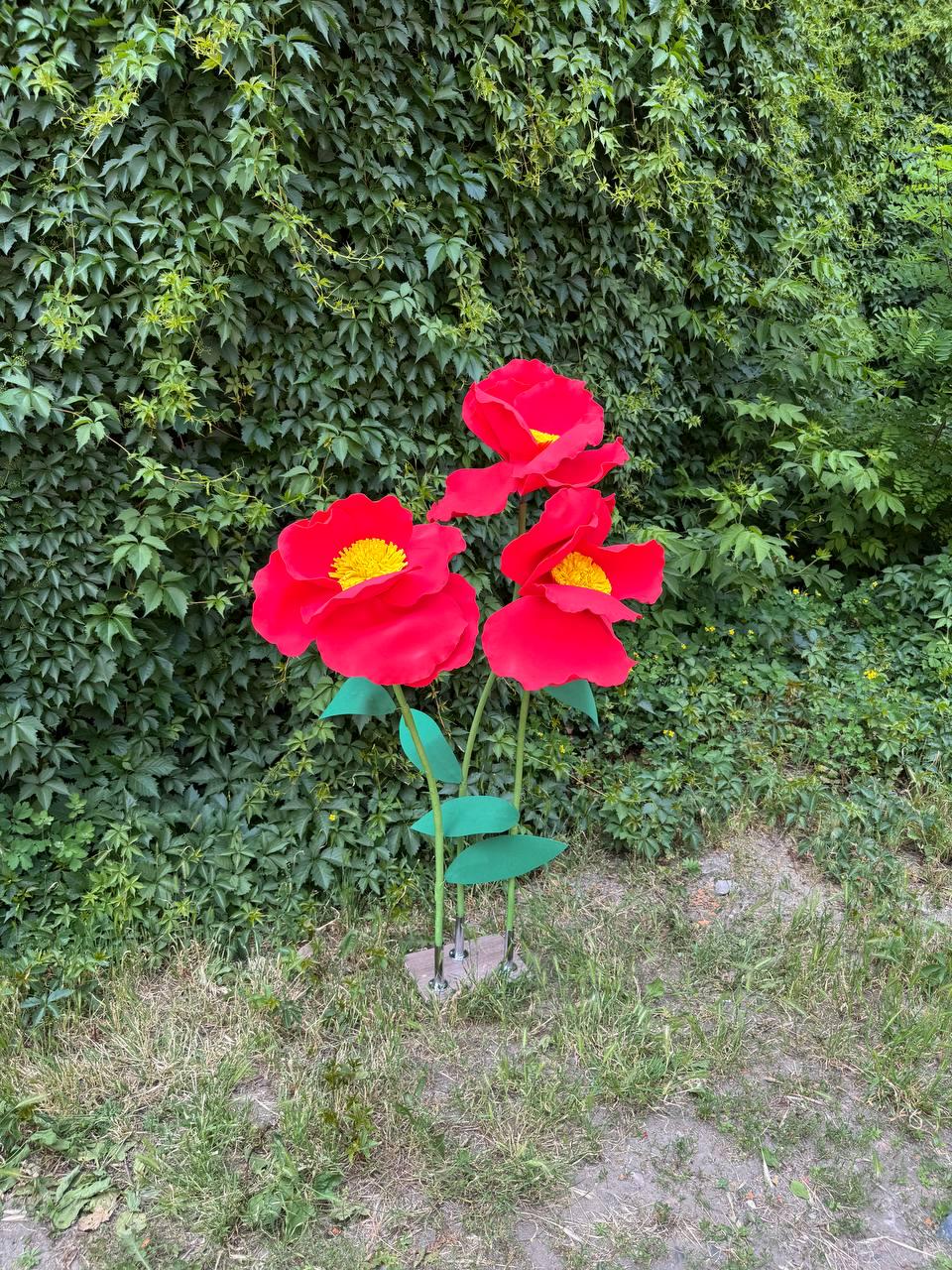 May include: Three large, artificial red flowers with bright yellow centers, mounted on thin green stems with green leaves. The flowers are attached to a small, rectangular base, creating a decorative display of oversized, vibrant red poppy-like blooms.