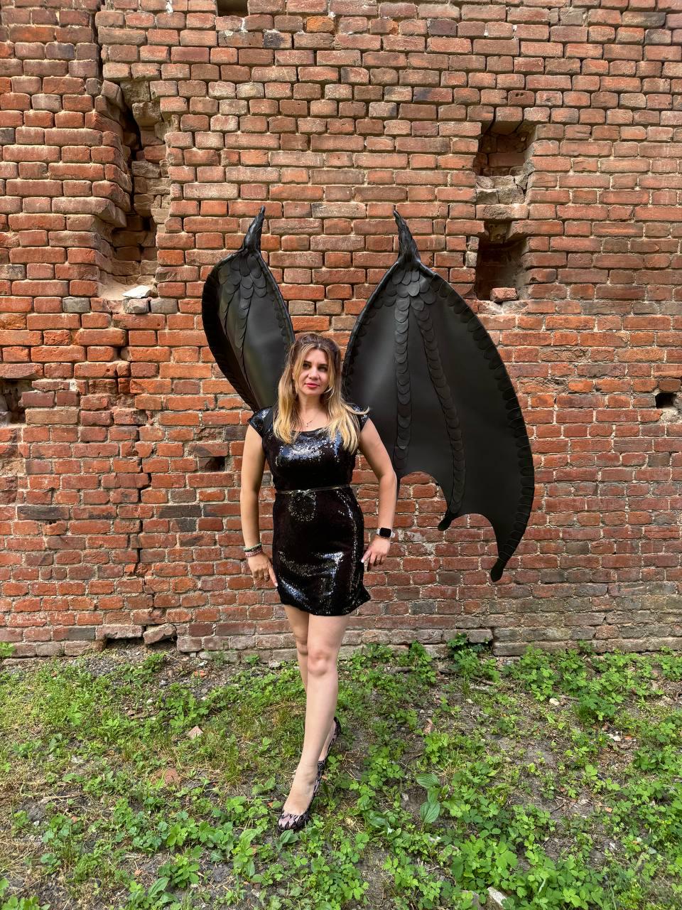 May include: A black sequined mini dress with a round neckline and short sleeves, paired with large, structured black dragon wings featuring a layered, scale-like texture. The wings are tall, pointed, and extend upwards behind the shoulders.