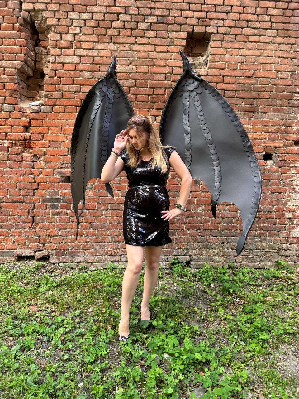 May include: A black sequined mini dress paired with large, articulated black dragon wings featuring a layered scale pattern. The wings are tall and pointed, extending upwards from the back, while the dress is sleeveless with a fitted silhouette.