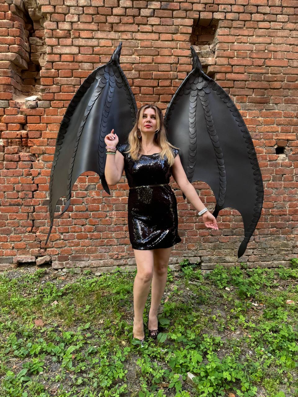 May include: A black sequined mini dress paired with large, articulated black dragon wings featuring a scale-like texture. The wings are tall and pointed, extending upwards from the back, while the dress has a scoop neckline and short sleeves.