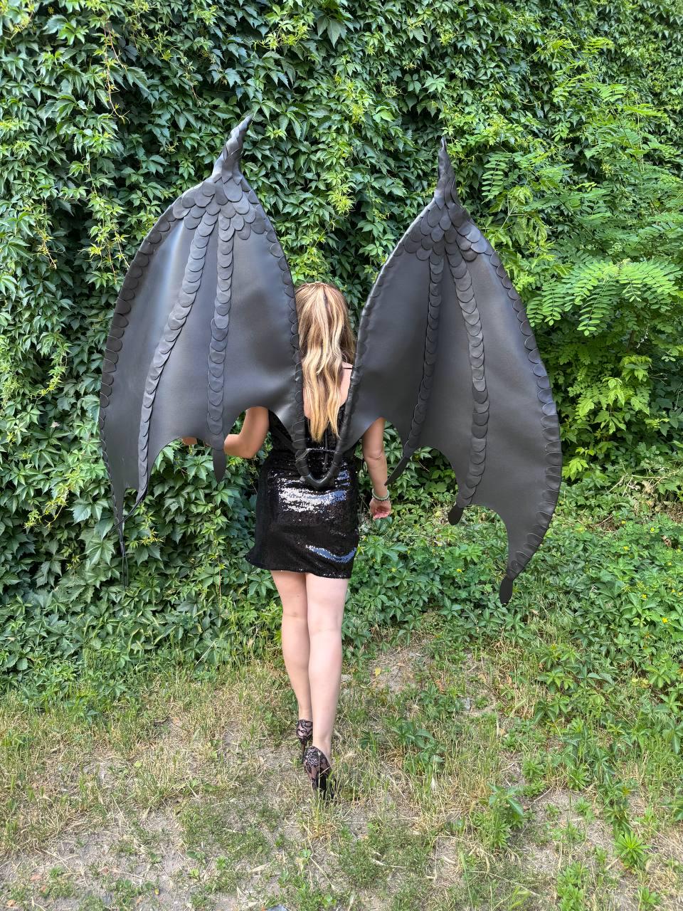 May include: Large, black, articulated dragon-style wings featuring a layered, scale-like texture along the structural ribs. The wings are mounted on a harness worn over a black, sleeveless, sequined mini dress with a straight hemline.