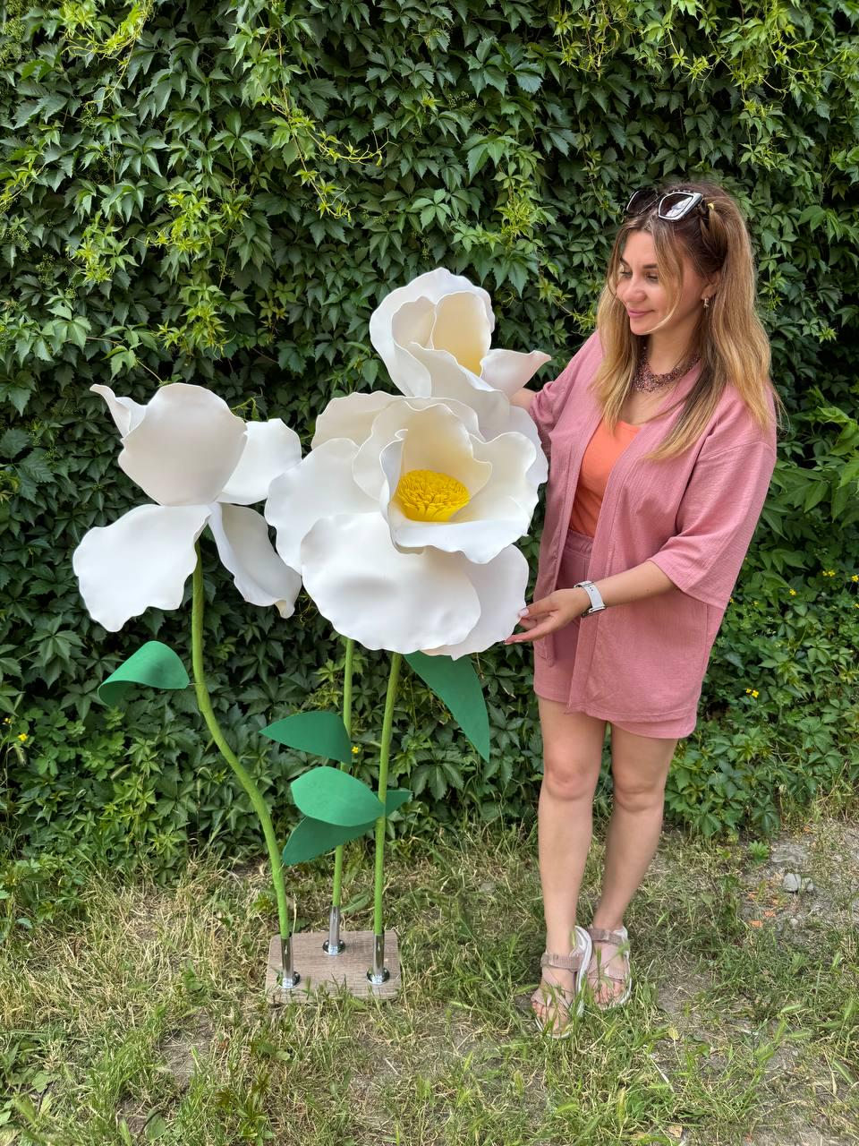 May include: Three oversized artificial white flowers with yellow centers, mounted on green stems with green leaves. The stems are attached to a rectangular wooden base with metal hardware. The flowers are large, sculptural, and white in color.