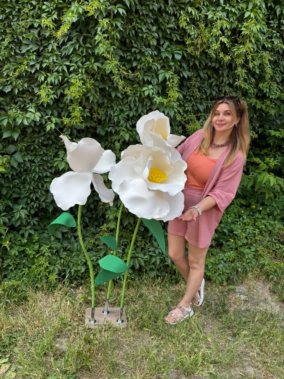 May include: Two large, oversized white artificial flowers with yellow centers, mounted on thin green stems with green leaves. The flowers are attached to a square wooden base, standing upright as a decorative display piece for indoor or outdoor use.